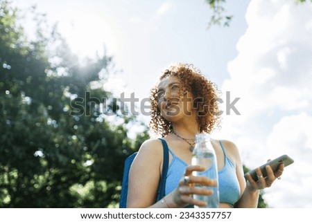 Low angle view of female in blue sport top on her way to gym for morning pilates training and yoga practice, holding smartphone and bottle of fresh water pictured on background of sunny and cloudy sky