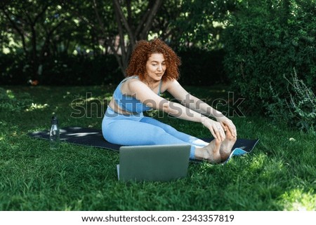 Horizontal portrait of ginger head female practicing yoga online outdoor, sitting in front of laptop on mat in park with bottle of fresh water behind her, stretching legs, touching her toes Royalty-Free Stock Photo #2343357819