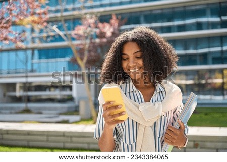 Smiling African American girl walking in university park using mobile phone shopping online or texting messages. Happy female student holding smartphone chatting standing in campus area.