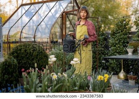 Portrait of a young woman as florist taking care of flowers and plants in garden with vintage greenhouse on background. Concept of hobby and gardening Royalty-Free Stock Photo #2343387583