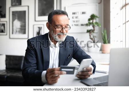 Cheerful caucasian senior businessman in suit, glasses pay at phone at table with computer, uses credit card in modern office interior. Finance, work remotely, business banking app, online shopping