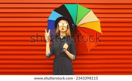 Autumn portrait of stylish young woman posing with colorful umbrella wearing black round hat blowing her lips sends kiss on red background