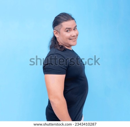 A smiling biracial male facing sideways to the right with head turned forwards while hands are inside his pockets. Isolated on a blue background.