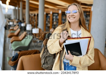 Happy smiling cute pretty blond girl, positive female teenage high school student holding backpack looking at camera standing in modern university or college campus library, portrait.