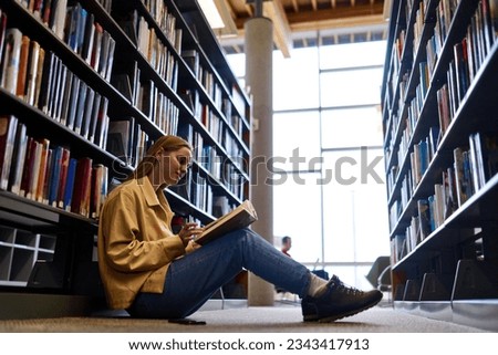 Smart pretty creative girl student holding book sitting on floor among bookshelves in modern university campus library or bookstore thinking of college course study reading literature doing research.