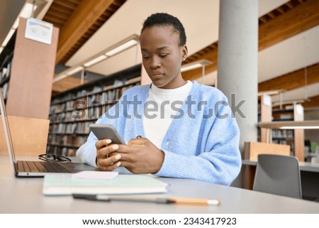 African girl student using mobile cell phone and laptop sitting at desk in university college library. Female Black learner holding cellphone elearning on computer tech in university campus. Royalty-Free Stock Photo #2343417923
