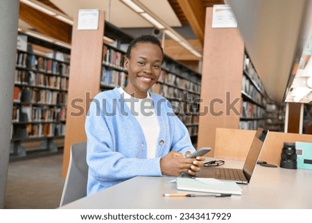 Happy African Black girl student using mobile phone and laptop sitting at desk in university college library. Female Black learner holding cellphone elearning on computer tech in campus. Portrait Royalty-Free Stock Photo #2343417929