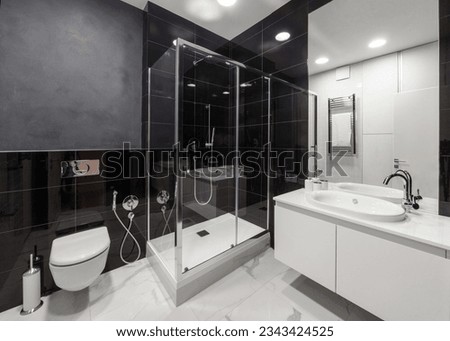 spacious bathroom with black tile on wall and white on floor, shower cabin with glass doors, modern washbowl with faucet and wc toilet with closed lid Royalty-Free Stock Photo #2343424525