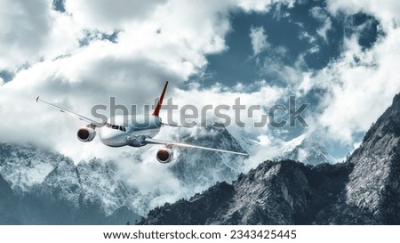 Airplane is flying over low clouds against mountains with snowy peaks. Landscape. Passenger airplane, cloudy sky, rocks, snow. Passenger aircraft. Business, commercial travel. Aerial view of plane