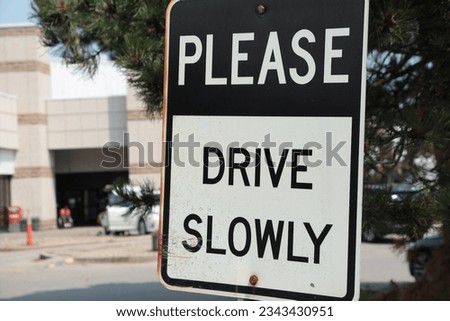 please drive slowly caption writing text rectangle metal sign with parking lot behind, white and black capital letters with white and black background