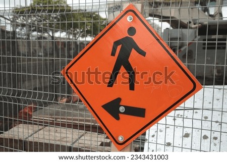diamond orange sign with arrow pointing left and illustration of person walking right in black print fastened to rectangle silver fence closed construction area with traffic passing frame left, angle