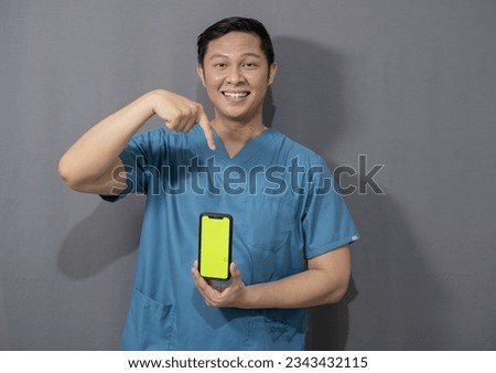Happy asian man working as medical worker wearing medical suit holding mobile phone, showing green screen, promoting