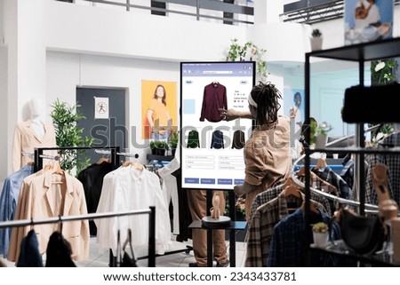 Client using touch screen board in trendy clothing store, shopping for fashion collection items in shop. Young buyer buying clothes on interactive monitor, self ordering concept. Royalty-Free Stock Photo #2343433781