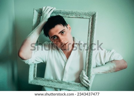 A young man sits beside a white wall, his hands covered in white paint as he holds an empty frame from a painting. The artist and his art, breaking through boundaries and thinking outside the box.