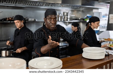 Skilled man executive chef standing in modern restaurant kitchen and gesturing emotionally behind while his team working Royalty-Free Stock Photo #2343442345