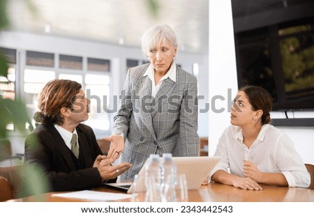 Portrait of strict senior female boss conducting work meeting with subordinates, standing near table and talking to man Royalty-Free Stock Photo #2343442543