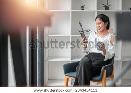 Cheerful millennial Asian woman using mobile phone, chatting on web, working or studying online, sitting in armchair against white wall, copy space.