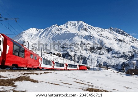 On a sunny winter day, tourists ride in a Glacier Express train and, thru the panoramic windows, enjoy the view of snowy Rossbodenstock mountain under blue clear sky, in Andermatt, Uri, Switzerland Royalty-Free Stock Photo #2343456711