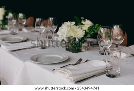 view of a table set for a party with a bouquet of white flowers and wine glasses next to the plates and tablecloth Royalty-Free Stock Photo #2343494741