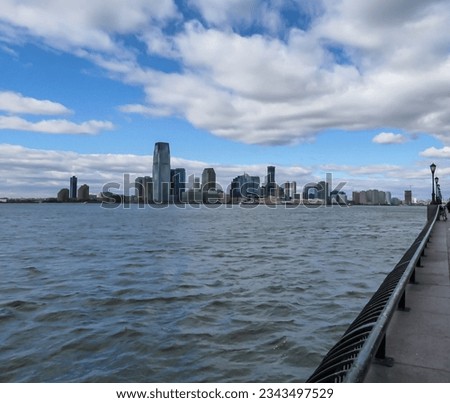 Breath-taking view on Downtown New York City skyline and Hudson River seen from the Battery Park. There is a long promenade along the river. Few clouds above the city. Tall skyscrapers. Royalty-Free Stock Photo #2343497529