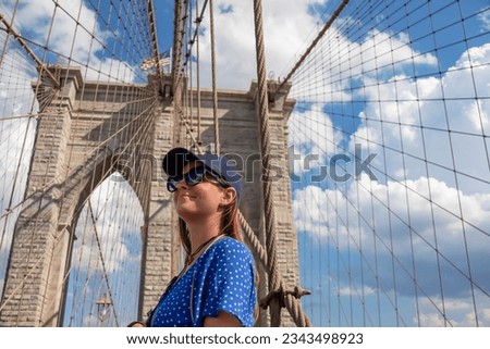 Woman in sunglasses and a cap standing in from of a gate of Brooklyn Bridge with a waving American flag on top of it contrasted with a blue sky with puffy, white clouds. Suspension bridge in New York