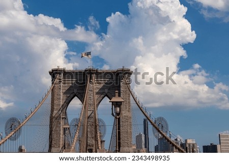The gate of Brooklyn Bridge with a waving American flag on top of it contrasted with a blue sky with puffy, white clouds. Suspension bridge in New York City. Numerous skyscrapers in the back.