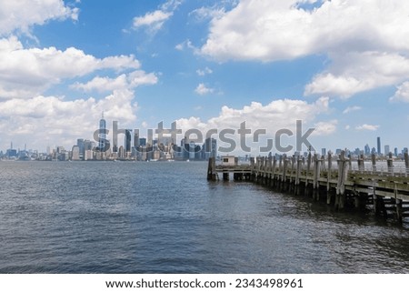 A wooden pier on the Hudson River with captivating New York urban skyline with striking and modern skyscrapers in the back, seen from The Battery Park. Think clouds above the city. Modern city. Royalty-Free Stock Photo #2343498961