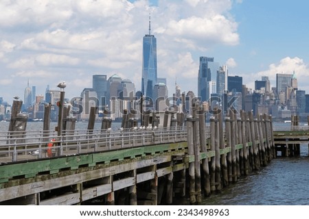 A wooden pier on the Hudson River with captivating New York urban skyline with striking and modern skyscrapers in the back, seen from The Battery Park. Think clouds above the city. Modern city. Royalty-Free Stock Photo #2343498963