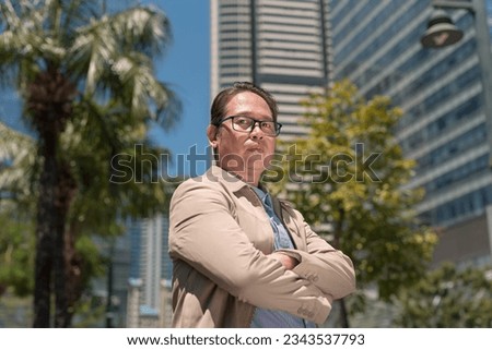 An unamused middle aged asian man with arms crossed standing by the sidewalk in the city. Royalty-Free Stock Photo #2343537793