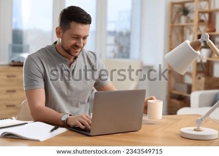 Happy man with cup of drink working on laptop at wooden desk in room