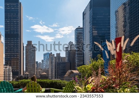 A woman sitting at the rooftop with the city skyline view and modern skyscrapers against a sunny blue sky in New York. She is enjoying the sunny day. Relaxing and recharging. Some greenery on the side