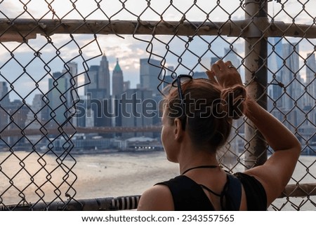 A woman with sunglasses on her head looking through a hole in a fence above Hudson River on a panoramic view of New York City's skyscrapers. Brooklyn Bridge in front. Overcast. Missing freedom