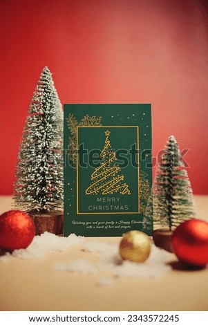 Selective focus of greeting card, artificial pine tree, mini ball and fake snow for Christmas decoration. Red and blurry background.