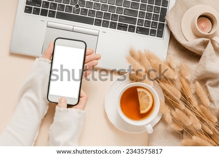 Woman hold mobile phone with white screen, laptop background with tea, candle, lagurus grass on linen cloth, aesthetic home workplace, cozy feminine office desk. Website promotion, business branding Royalty-Free Stock Photo #2343575817