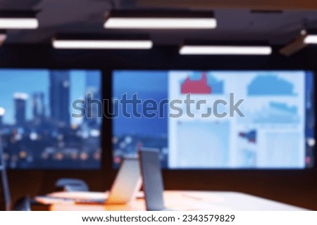 Blurred office background. Modern Empty Meeting Room with Big Conference Table with Various Documents and Laptop on it. Wall TV Showing Company Statistics, Graphs. Late Evening or Night City Outside.