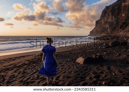 Woman in dress walking barefoot on volcanic sand beach Playa del Ingles during sunset in Valle Gran Rey on La Gomera, Canary Islands, Spain, Europe. Calm atmosphere seaside. Massive cliffs La Mercia Royalty-Free Stock Photo #2343585311