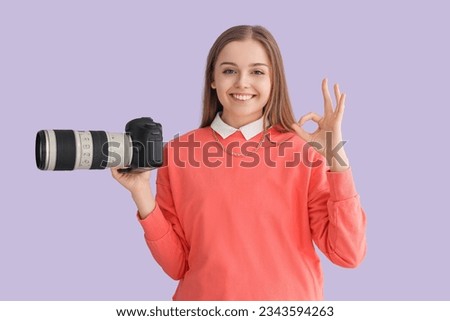 Young female photographer with professional camera showing OK on lilac background