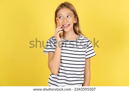 Pleasant looking happy Caucasian kid girl wearing striped T-shirt over yellow background has nice telephone conversation and looks aside, has nice mood and smiles positively while talks via cell phone
