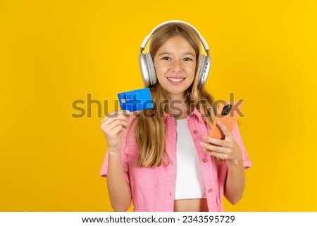 Caucasian kid girl wearing pink dress over yellow background wears stereo headphones on ears holds modern mobile phone and credit card
