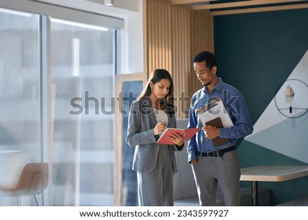 Two multicultural workers discussing corporate work plan standing in office. Busy international business team coworkers talking on marketing project sharing ideas taking notes working together.