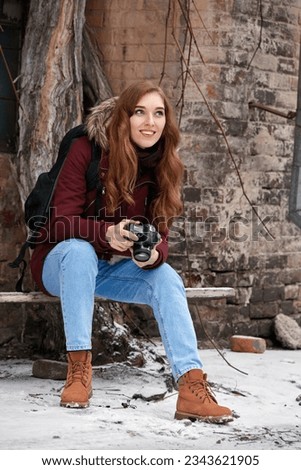 Happy attractive redhead girl with camera sitting on bench outdoors in winter city
