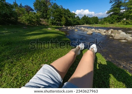 Close-up of the legs of a woman in denim shorts and white sports shoes sitting on the green grass by a mountain river on a sunny day