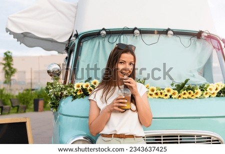 Young woman buying street food from a food truck at park. Caucasian girl holding a cup of lemonade to go at vintage food truck. Street food business, festival. Mobile cafe. Coffee to go