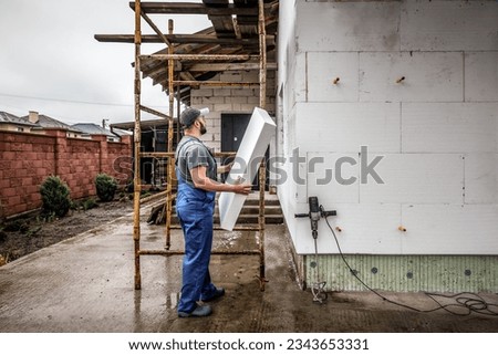 Professional master holds adhesive styrofoam to the wall. Building insulation saves energy. Facade master. Industrial theme Royalty-Free Stock Photo #2343653331