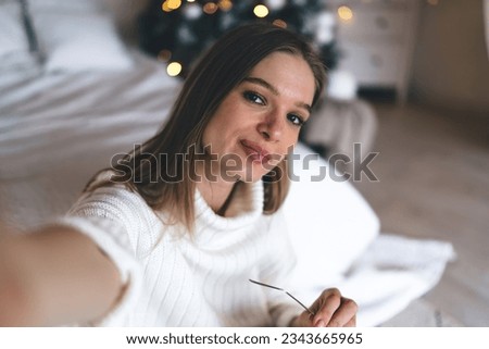From above portrait of confident young woman in warm clothes looking at camera and taking selfie in bedroom during Christmas celebration
