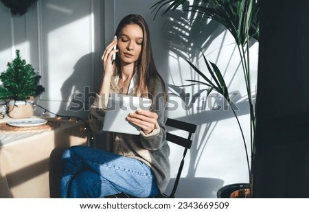 Confident young female in casual clothes sitting at table with shades of sunlight and attending call on mobile phone while reading cover page of literary book in hand in daytime at home Royalty-Free Stock Photo #2343669509