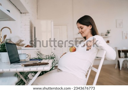 Side view of positive pregnant woman in white clothes jokingly placing apple on belly while sitting at table with laptop in light spacious room