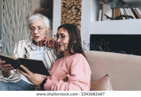 Positive elderly diverse women in eyeglasses sitting in living room watching photo album together enjoying pleasant memories having great time