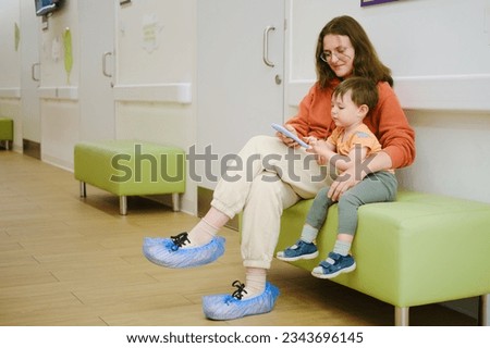 The baby and his mother are sitting together in the hospital hall, playing a game on their phone to pass the time before their doctor's visit. Kid boy aged two years (two-year-old)