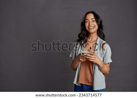 Happy casual beautiful woman using smartphone isolated against grey background. Smiling hispanic young woman with backpack using mobile phone isolated against gray background with copy space. Royalty-Free Stock Photo #2343714577